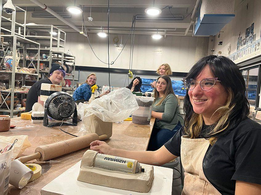 Seven students smiling for the camera as they are also making molds and slip casting in the ceramics classroom with view of the storage shelving and windows in the background.