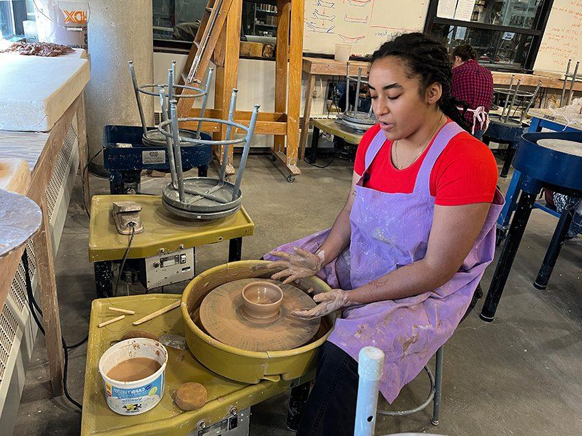 Student sitting at a potters wheel throwing a small bowl with a view of additional potter’s wheels in the background in the ceramics classroom.